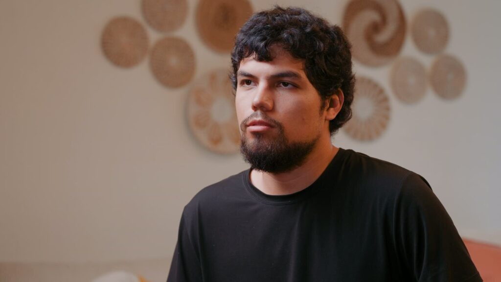 A young man speaking on camera in a softly lit living room, from the Corazón de Vida testimonial film