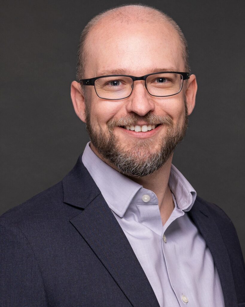 Corporate headshot of a smiling man with glasses in a navy blazer
