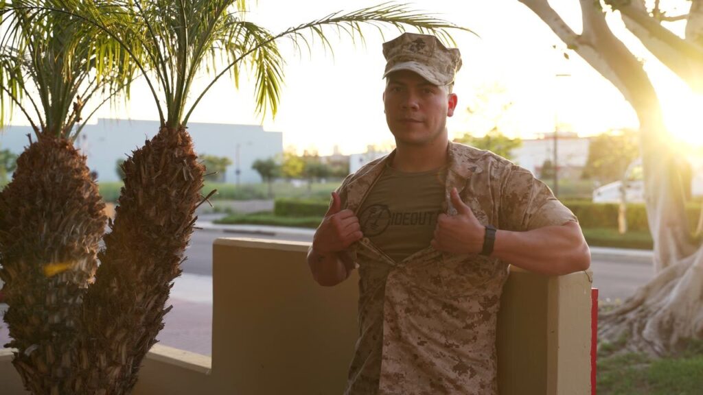 A Marine in desert camouflage opening his uniform to show a Hideout Fitness shirt at golden hour
