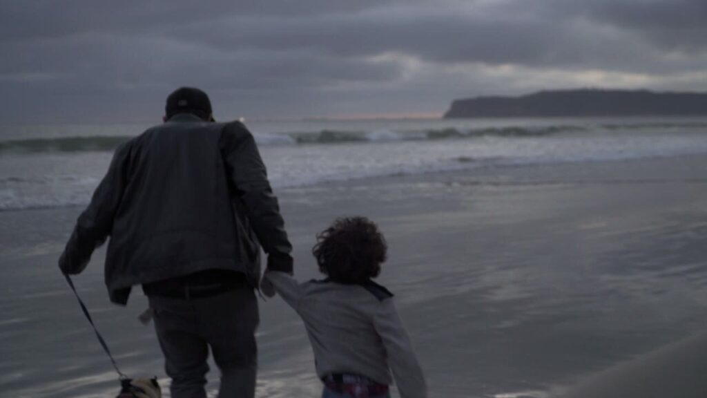 Eddie walking his son hand in hand to the surf at Coronado Dog Beach at dusk, the dog on a leash, seen from behind