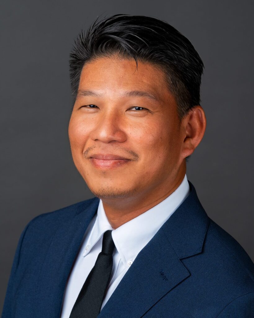 Team headshot of a man in a navy suit and black tie, shot at his office