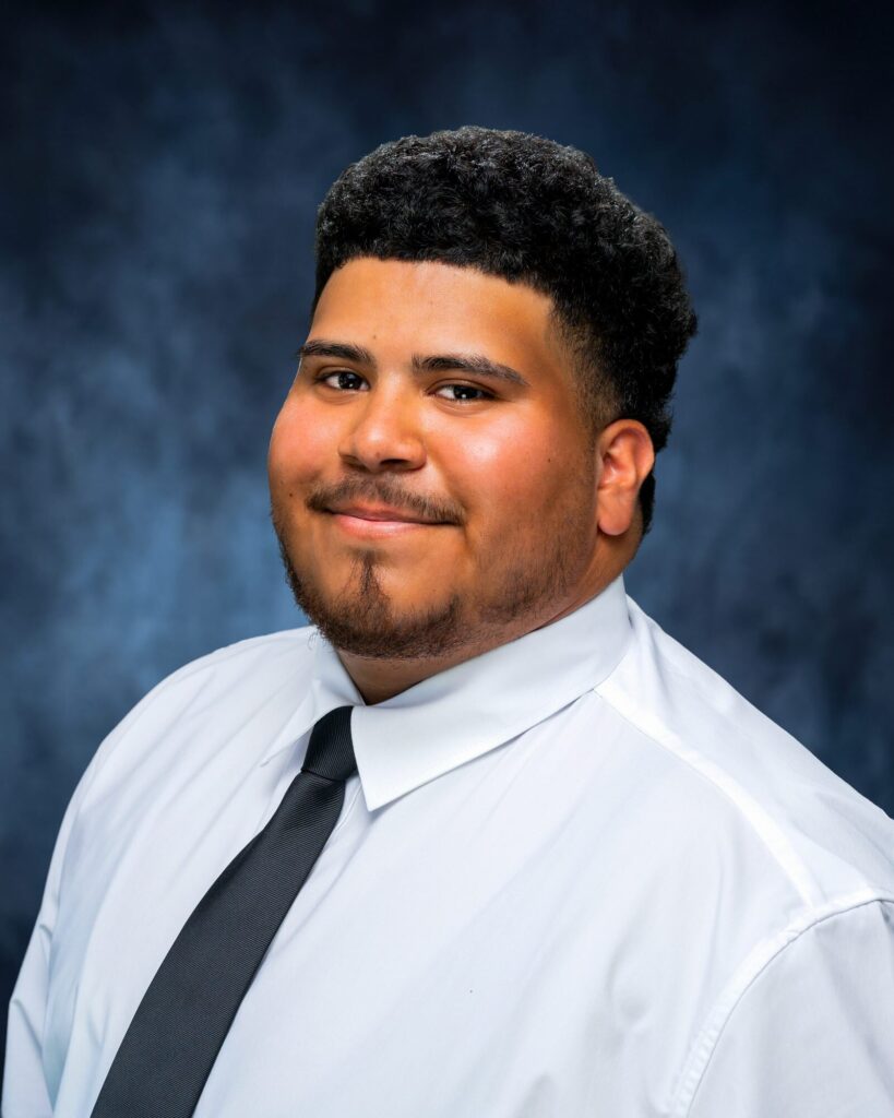 Team headshot of a young man in a white shirt and black tie on a blue backdrop