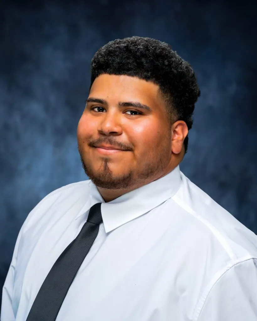 Team headshot of a young man in a white shirt and black tie on a blue backdrop